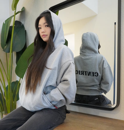 Person wearing a gray hoodie with a mirror reflection on a white surface with green plants.